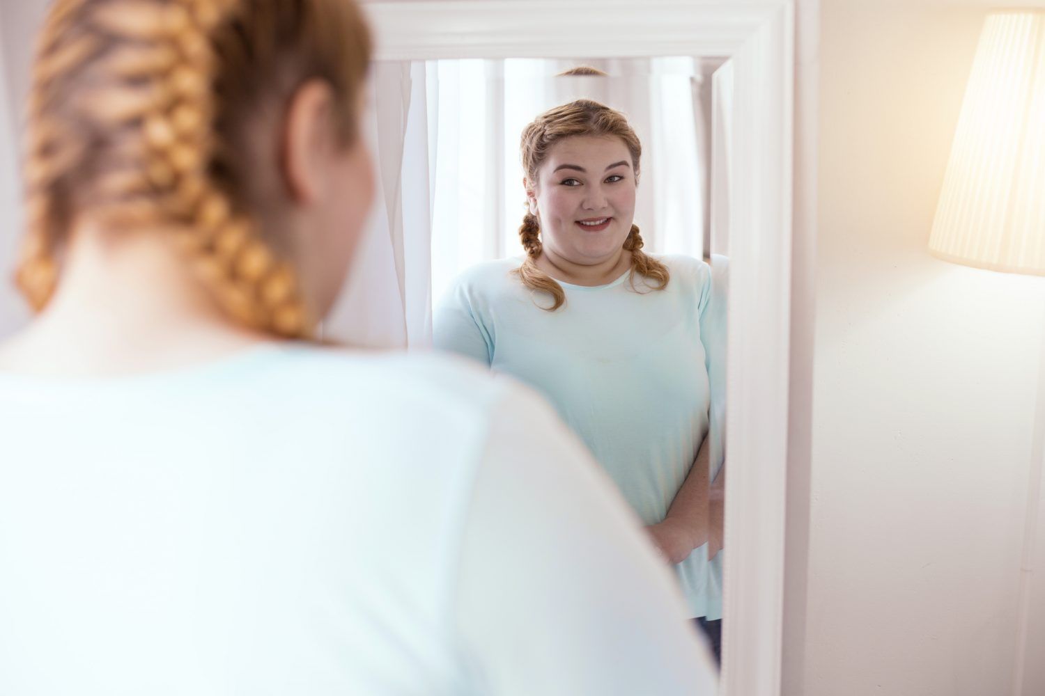 Junge Frau mit Zöpfen steht in einem hellen Raum vor einem Spiegel und schaut sich selbst an. Sie trägt ein hellblaues Oberteil und lächelt leicht. Die Spiegelung zeigt ihr Gesicht frontal, während sie von hinten nur teilweise zu sehen ist.