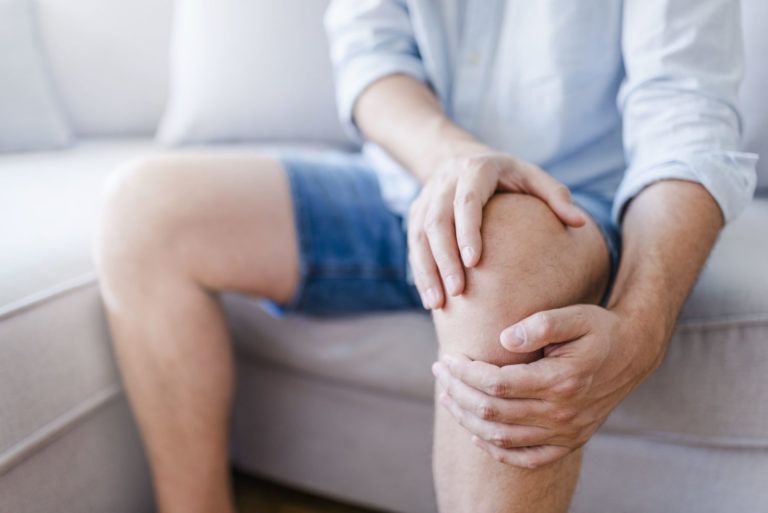 Person sitzt auf Sofa und hält schmerzhaft das rechte Knie mit beiden Händen