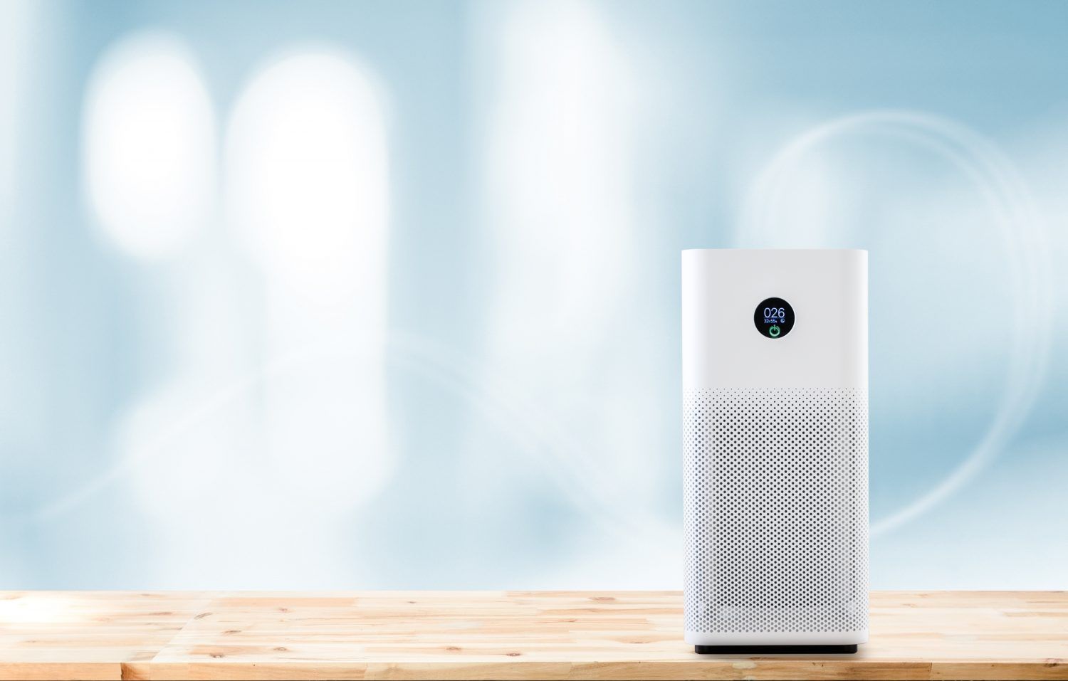 Weißer Luftreiniger mit digitalem Display steht auf einer hellen Holzoberfläche vor einem weich verschwommenen, hellblauen Hintergrund. Das Display zeigt Werte zur Luftqualität an.