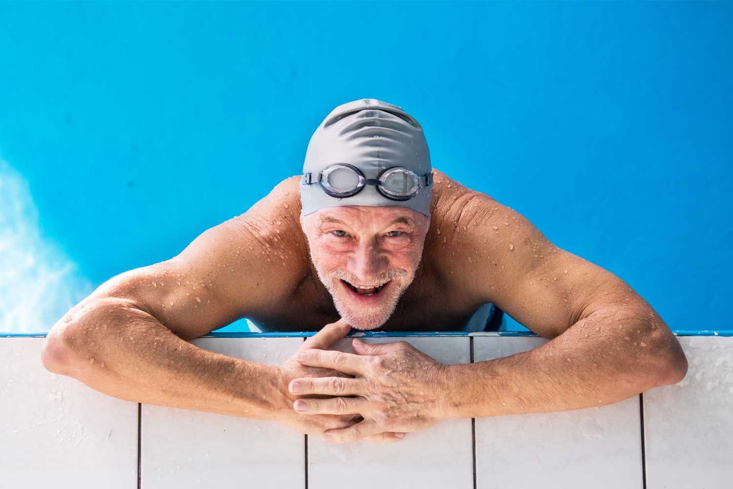 Älterer Mann mit Badekappe und Schwimmbrille lehnt lächelnd am Beckenrand eines Schwimmbads. Er ist oberkörperfrei, nass und wirkt fit und aktiv. Der Hintergrund zeigt klares, blaues Wasser.