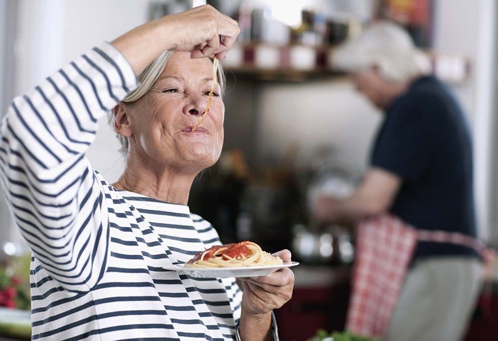 Ältere Frau genießt einen Teller Spaghetti: Im Vordergrund isst eine lächelnde Frau mit grauen Haaren genüsslich Spaghetti mit Tomatensauce