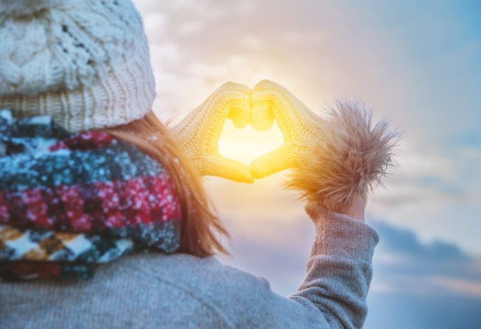 Person mit Winterkleidung formt mit Händen ein Herz vor der Sonne am Himmel
