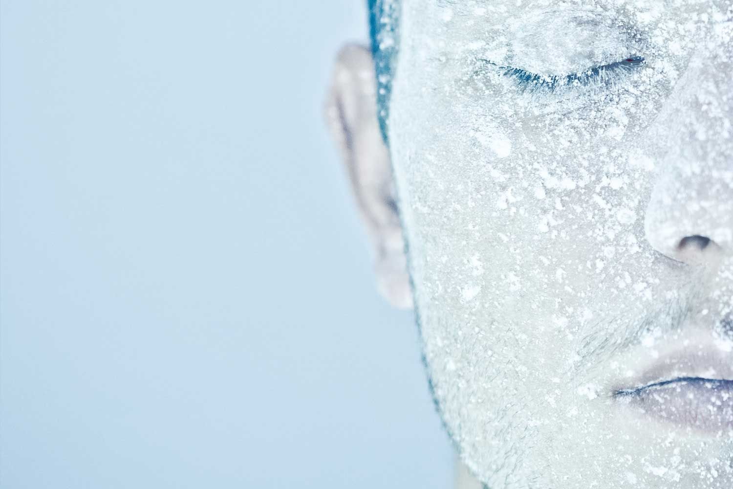 Nahaufnahme einer männlichen Gesichtshälfte mit geschlossenen Augen. Die Haut ist vollständig mit einer weißen Frost- oder Eisschicht bedeckt. Der Hintergrund ist hellblau, was die Kältewirkung unterstreicht.