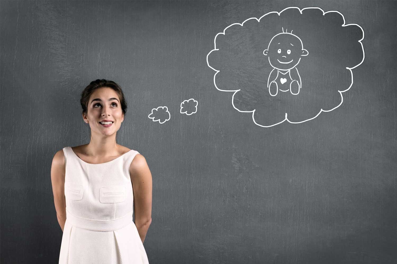 Junge Frau in weißem Kleid steht lächelnd vor einer dunklen Tafel und blickt nach oben. Auf die Tafel ist eine große Gedankenblase gezeichnet, die ein gezeichnetes Baby mit Herz auf dem Strampler zeigt. Zwei kleinere Wolken deuten auf den Übergang zur großen Gedankenblase hin.