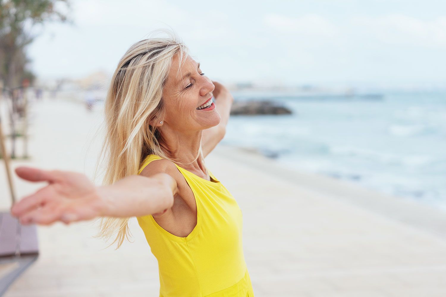 Ältere Frau mit langen, blonden Haaren trägt ein gelbes ärmelloses Kleid und steht mit ausgebreiteten Armen lachend an einer Strandpromenade. Der Wind weht ihr durch die Haare, im Hintergrund ist das Meer mit sanften Wellen zu sehen.