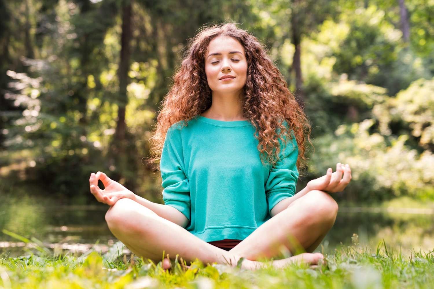 Junge Frau mit lockigem Haar sitzt barfuß im Schneidersitz auf einer Wiese und meditiert. Ihre Augen sind geschlossen, die Hände ruhen mit Daumen und Zeigefinger zusammengelegt auf den Knien. Im Hintergrund ist ein bewaldetes Gebiet mit einem Teich oder Bachlauf zu sehen.