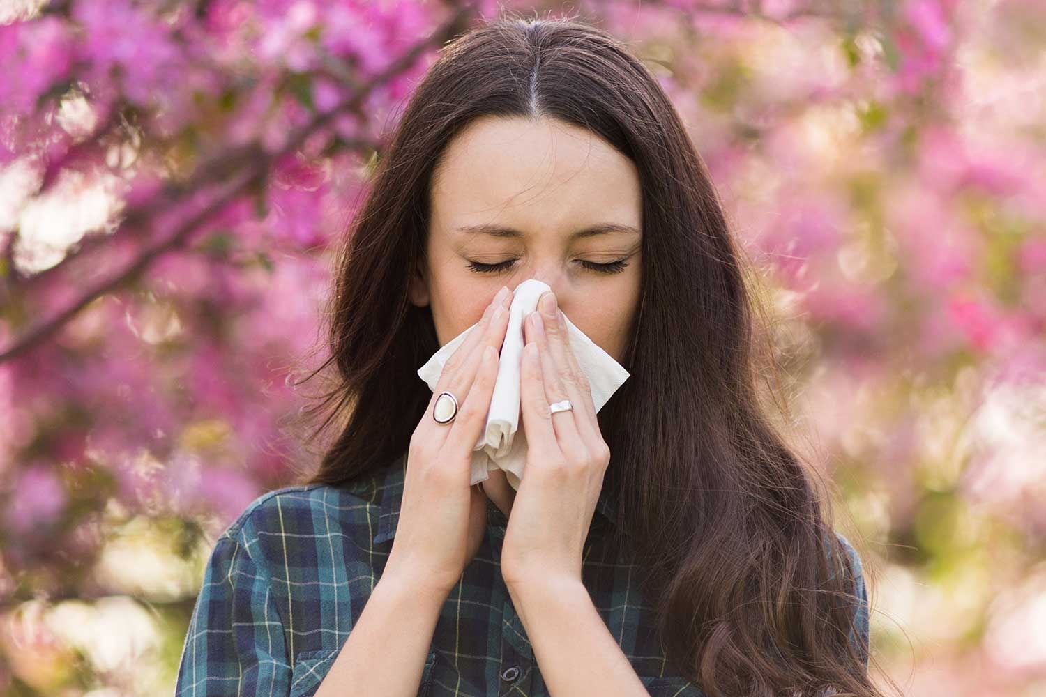 Junge Frau mit dunklen, langen Haaren steht vor einem blühenden Baum mit rosa Blüten und hält sich ein Taschentuch an die Nase. Ihre Augen sind geschlossen, und ihr Gesichtsausdruck deutet auf Niesen oder allergische Beschwerden hin.