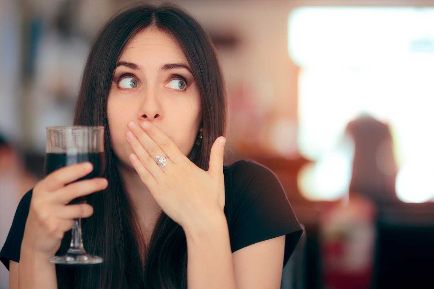 Junge Frau mit langen dunklen Haaren hält ein Glas mit dunkler Flüssigkeit in der einen Hand und bedeckt mit der anderen erschrocken ihren Mund. Sie blickt zur Seite und wirkt überrascht oder nachdenklich. Der Hintergrund ist unscharf und zeigt eine Café- oder Restaurantumgebung.