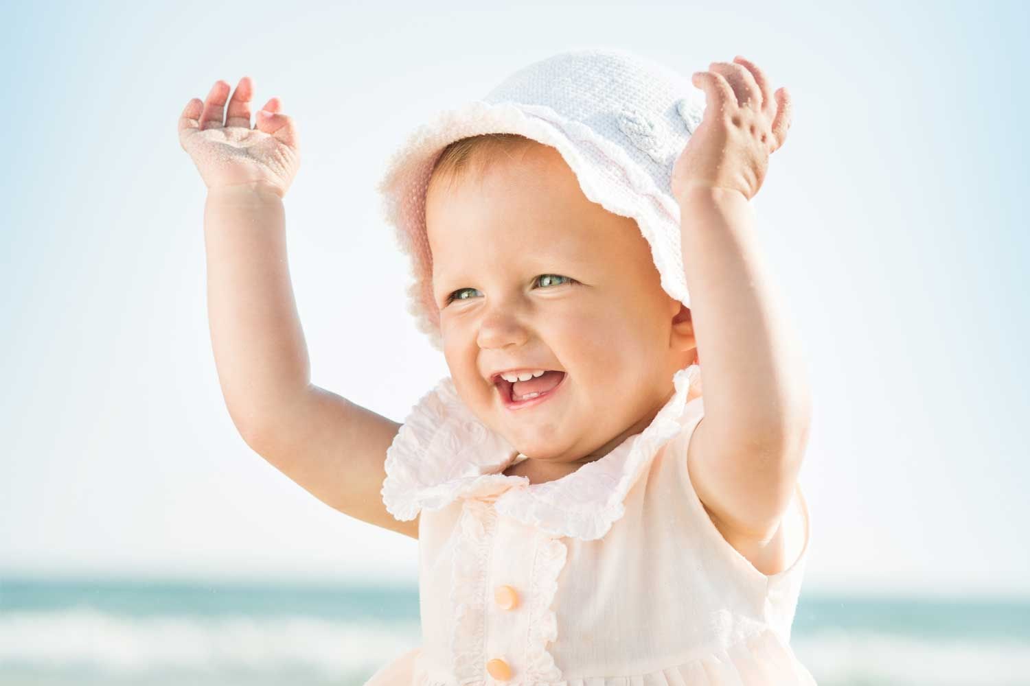 Fröhliches Kleinkind mit hellem Sonnenhut und ärmellosem Sommerkleid hebt beide Arme lachend in die Luft. Im Hintergrund ist verschwommen das Meer zu erkennen. Die Szene wirkt sonnig, unbeschwert und sommerlich.