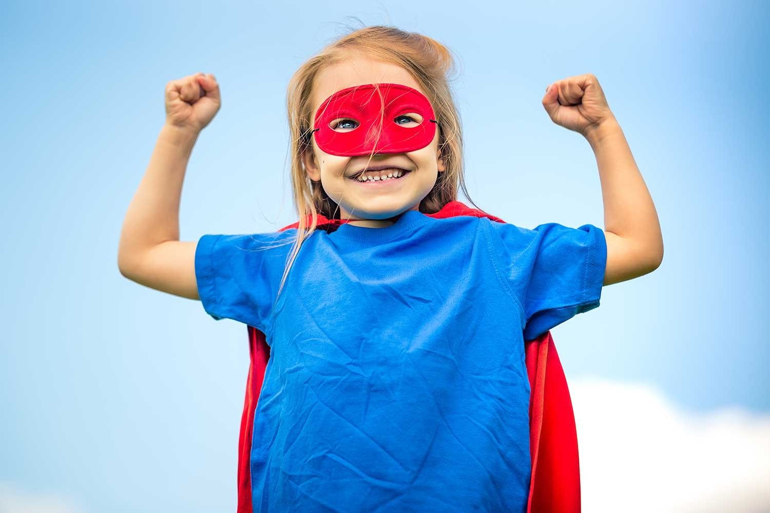 Lächelndes Mädchen trägt ein blaues T-Shirt, eine rote Maske und einen roten Umhang. Es hebt beide Arme in Siegerpose und schaut stolz in die Kamera. Der Himmel im Hintergrund ist blau mit vereinzelten Wolken.