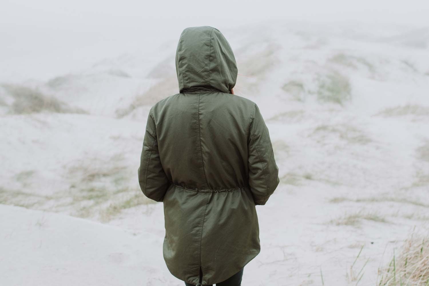 Person in Winterjacke mit Kapuze von hinten: Eine alleinstehende Person steht in einem nebligen Dünengebiet. Sie trägt eine olivgrüne Jacke mit Kapuze und blickt in die verschneite oder sandige Landschaft.
