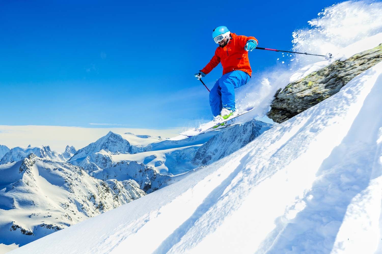 Skifahrer bei Abfahrt im Tiefschnee: Eine Person mit blauer Skihose, oranger Jacke und Helm fährt rasant eine verschneite Bergflanke hinab. Der Schnee wirbelt auf, im Hintergrund sind schneebedeckte Alpengipfel unter klarem, blauem Himmel zu sehen.